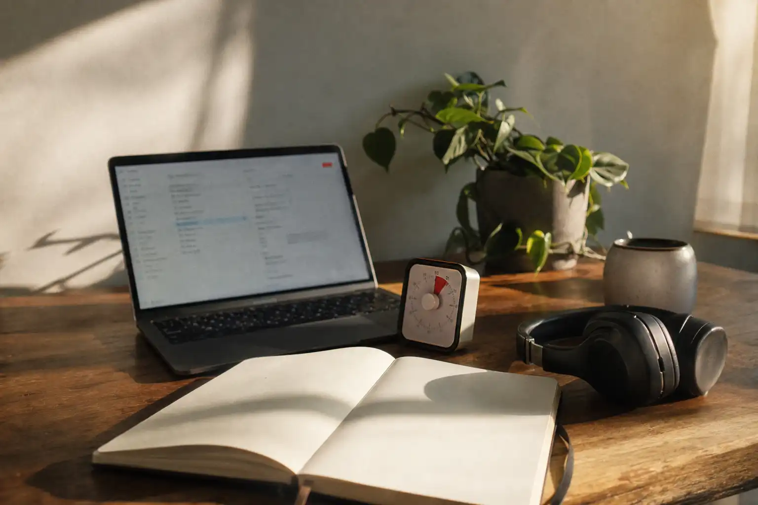 A quiet home workspace set up for a personal deep-work session: an open laptop with an unreadable blurred task interface, a paper notebook with blank-looking pages, a small analog timer, headphones resting beside a ceramic mug, and a single houseplant on a wooden desk near a window. No people, hands, feet, or readable text. Soft late-afternoon window light creates gentle shadows, with a slightly imperfect candid composition, subtle motion blur at the timer edge, fine digital noise, and natural lived-in desk details. Shot on Fujifilm X100V, 35mm, f/2, ISO 800.