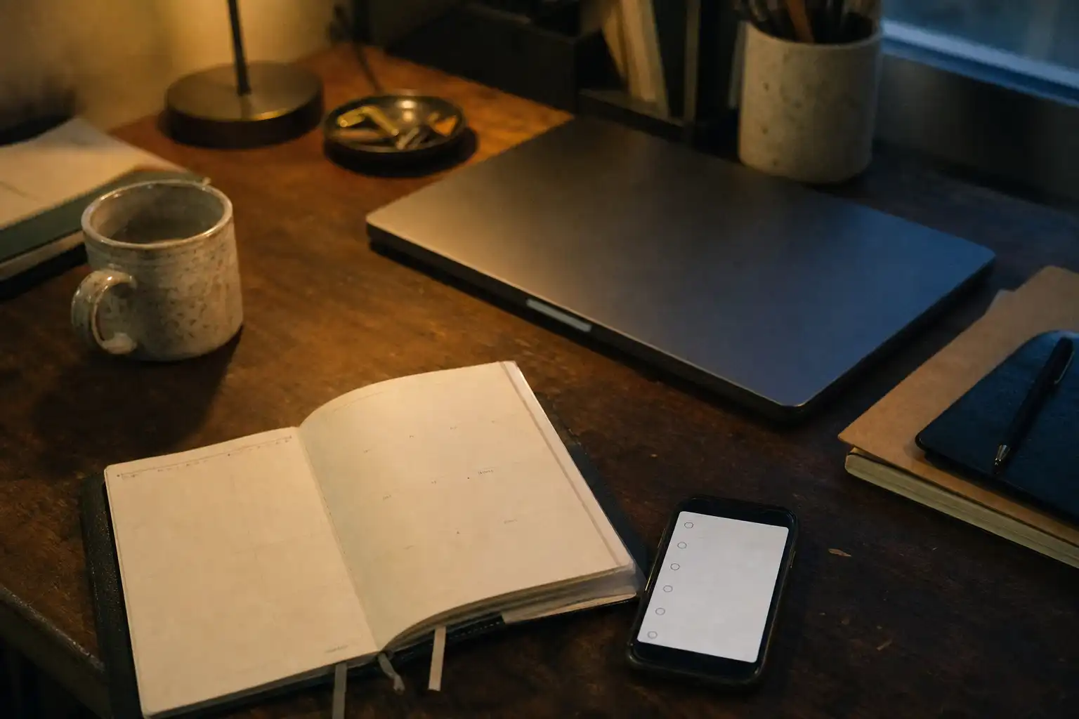 A candid evening photograph of a small smartphone resting beside an open paper planner and a closed laptop on a slightly cluttered wooden desk, the phone screen showing only simple abstract check circles with no readable text or logos, a ceramic mug and soft desk lamp in the background, overhead three-quarter composition, warm practical lamp light with cool window shadows, Fujifilm X100V, 35mm, f/2, ISO 800, slight motion blur, natural grain, and subtle low-light noise.