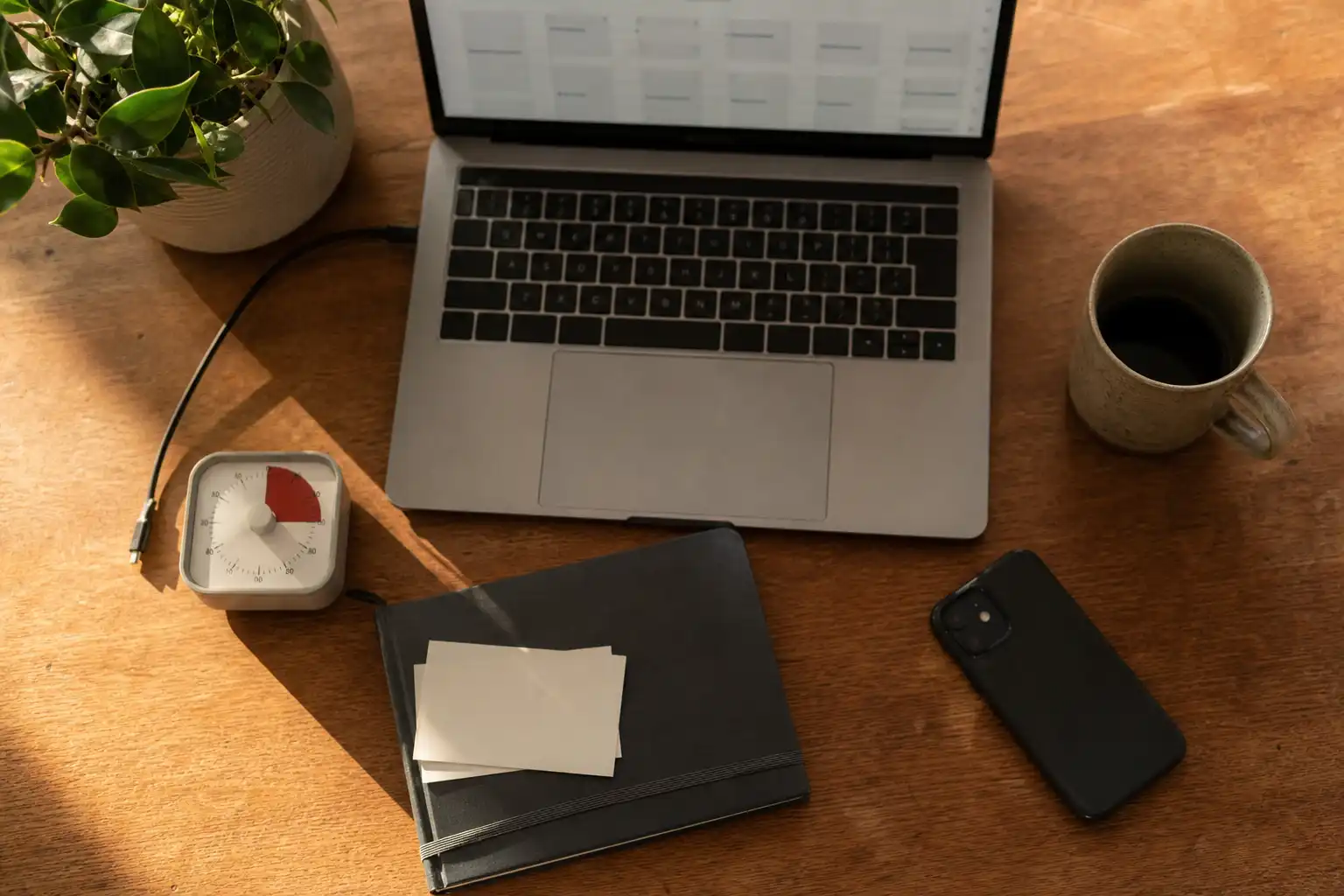 Editorial still-life photograph of a compact founder desk in late-afternoon window light: an open laptop with a softly blurred blank planning interface, a simple analog timer, an unmarked notebook with a few blank index cards, a phone face down, coffee mug, charging cable, and a small desk plant. Overhead three-quarter composition, realistic everyday clutter, calm but active atmosphere, warm natural lighting, no people and no readable text.
