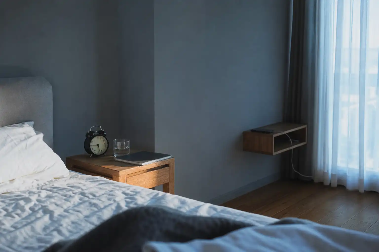 Editorial lifestyle photograph, no people and no text: a neatly made bed beside a small wooden nightstand in early blue morning light, with an analog alarm clock, a glass of water, a closed notebook, phone placed face-down on a charging shelf across the room, and soft daylight entering through partially open curtains. Calm realistic apartment bedroom, muted blue and warm wood palette, gentle shadows, wide horizontal composition with clear empty space for a blog header.